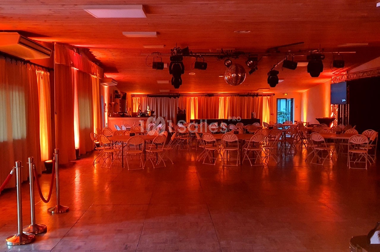Salle de réception avec tables et chaises pliantes, éclairée par des lumières chaudes et une boule disco au plafond.