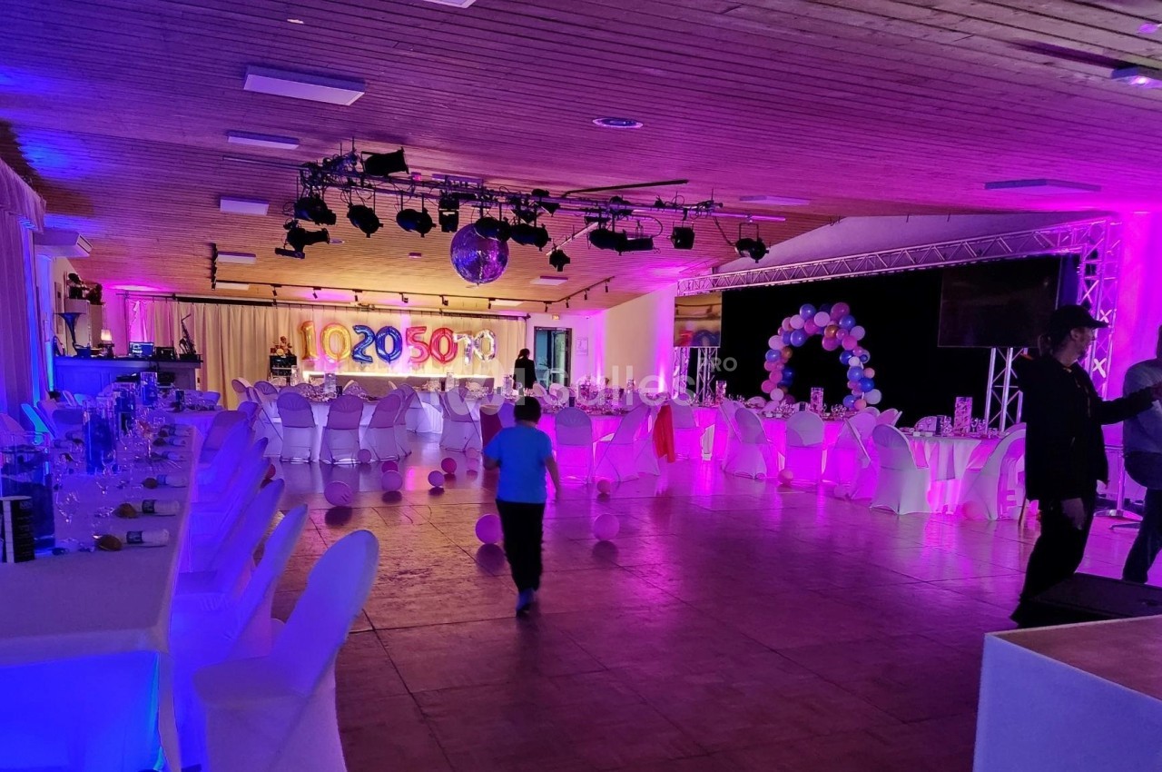 Salle de réception décorée avec des tables dressées, des ballons et un éclairage coloré, avec quelques personnes présentes.