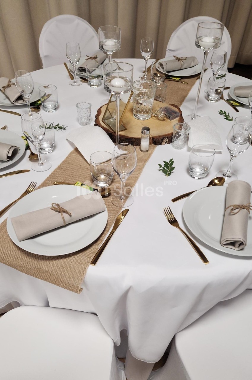 Table dressée avec une nappe blanche, couverts dorés, serviettes beige nouées, chemins de table en toile de jute et…