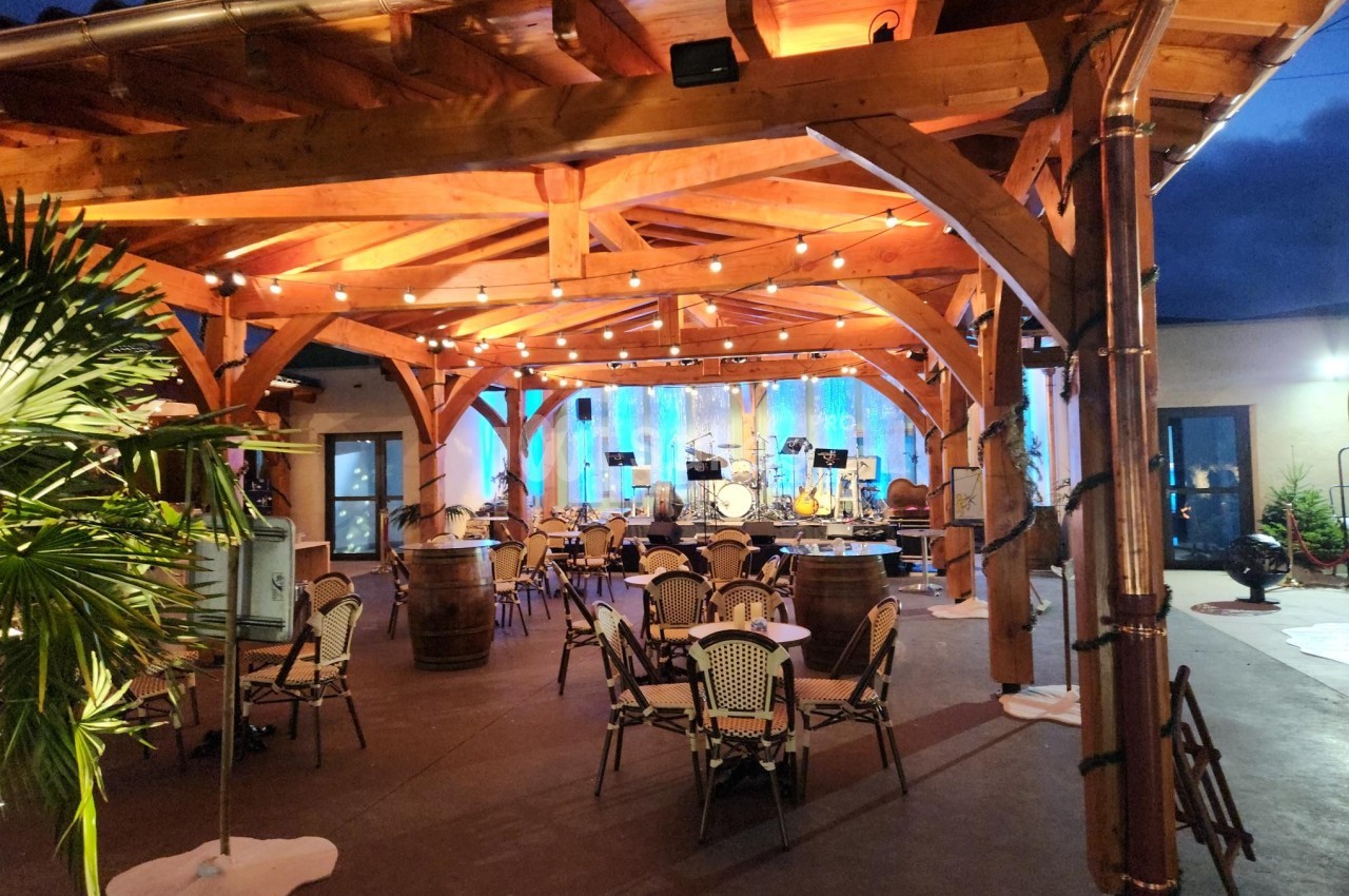 Structure en bois éclairée avec des guirlandes lumineuses, tables et chaises disposées pour un événement en soirée.