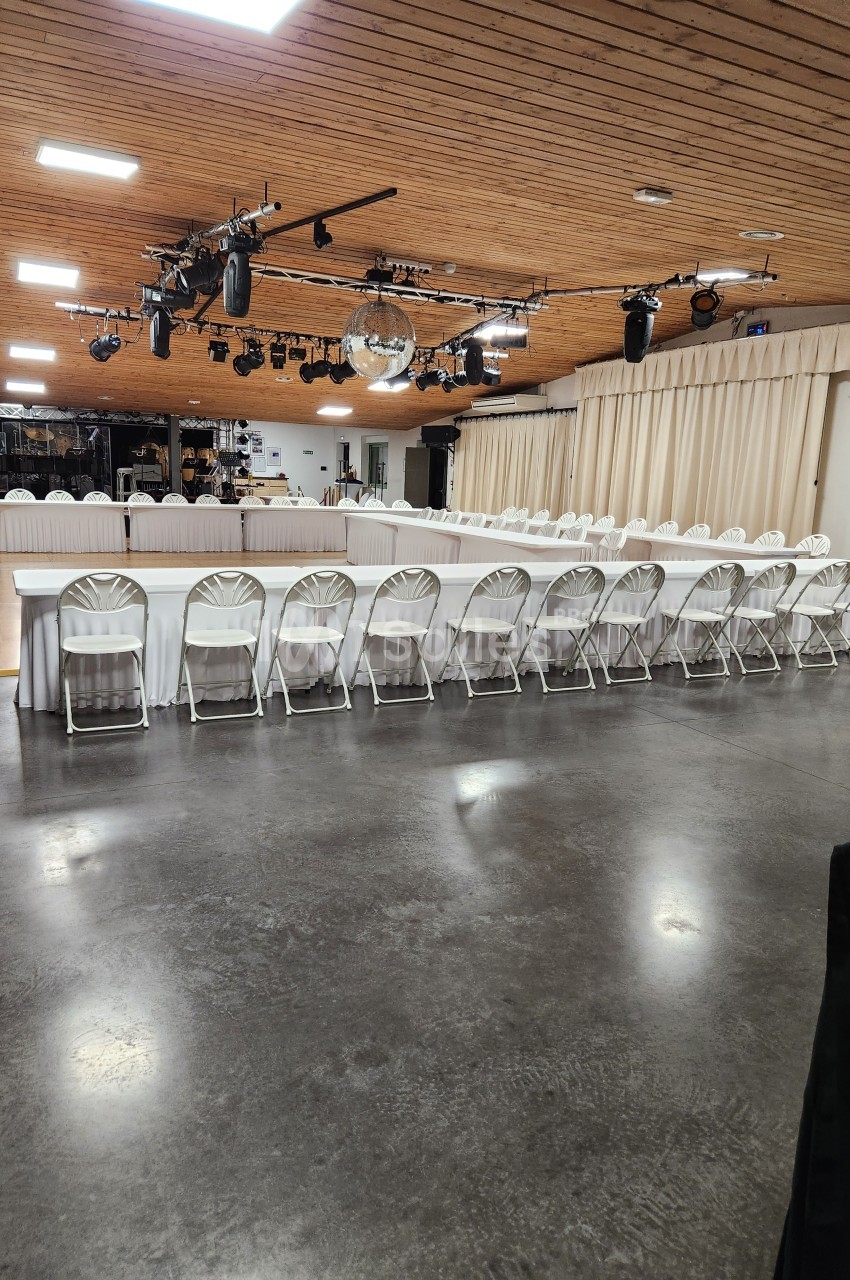 Salle avec un sol en béton, tables disposées en U avec nappes blanches, chaises pliantes et éclairage suspendu.