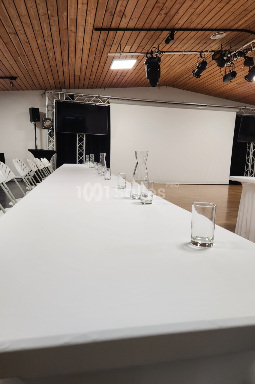 Salle de conférence avec une grande table blanche, des verres et des carafes, face à un écran de projection.