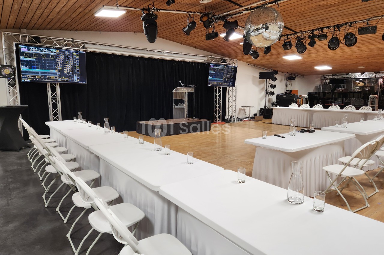Salle de conférence aménagée avec des tables blanches, des chaises, un pupitre et des écrans pour une présentation.