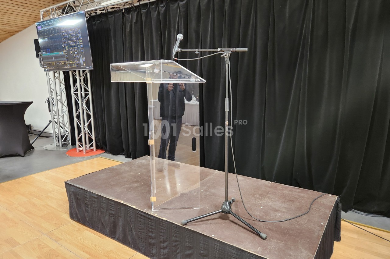 Un pupitre transparent avec microphone sur une petite estrade en bois, devant un rideau noir et un écran latéral.