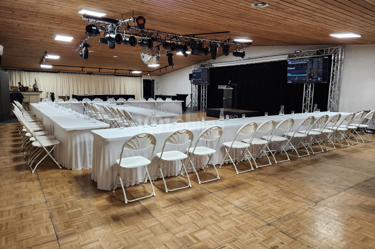 Salle de conférence avec tables disposées en U, chaises blanches pliantes et scène équipée de matériel audiovisuel.