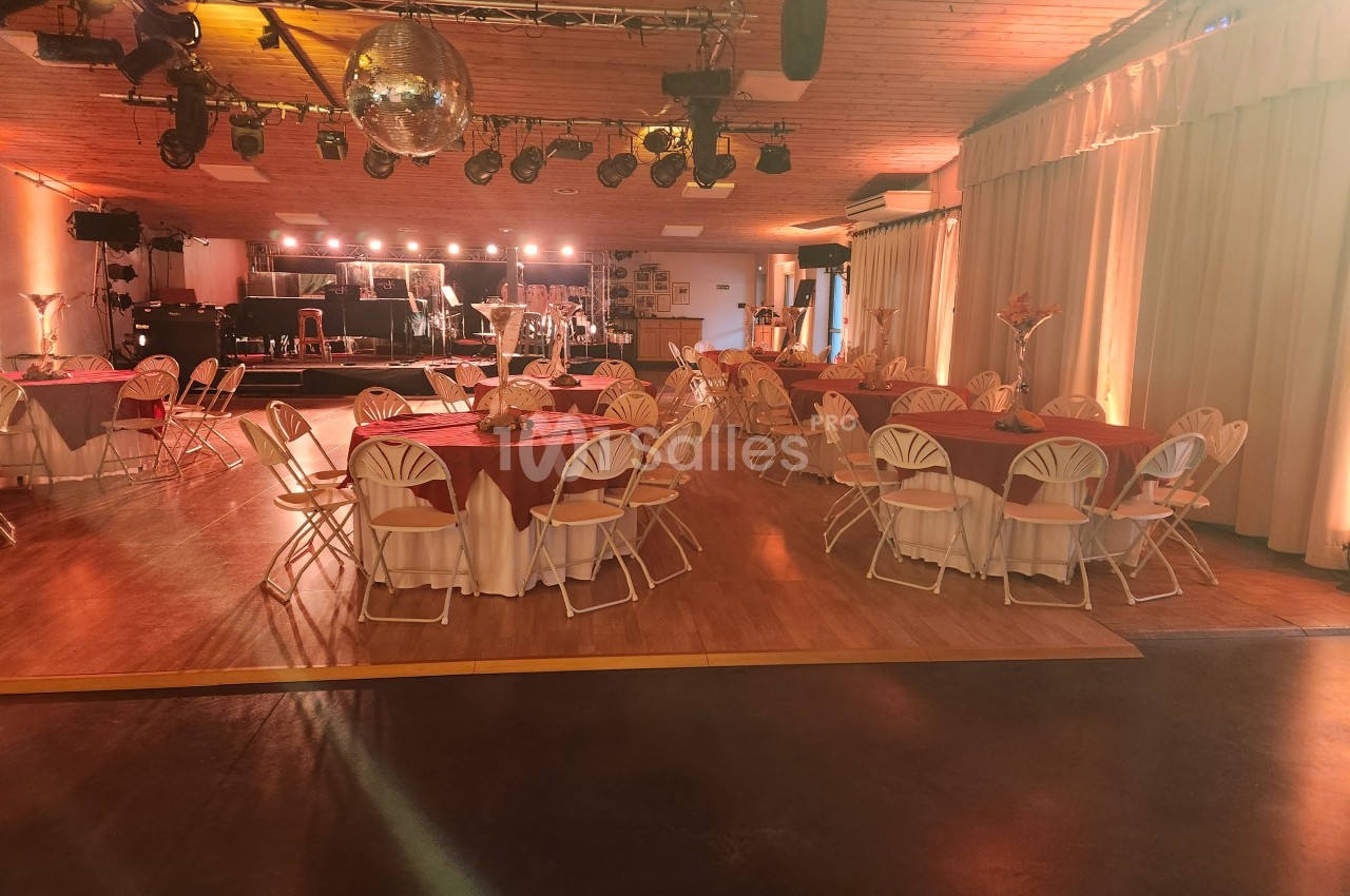 Salle de réception décorée avec des tables rondes nappées de rouge, éclairage chaleureux et scène en arrière-plan.