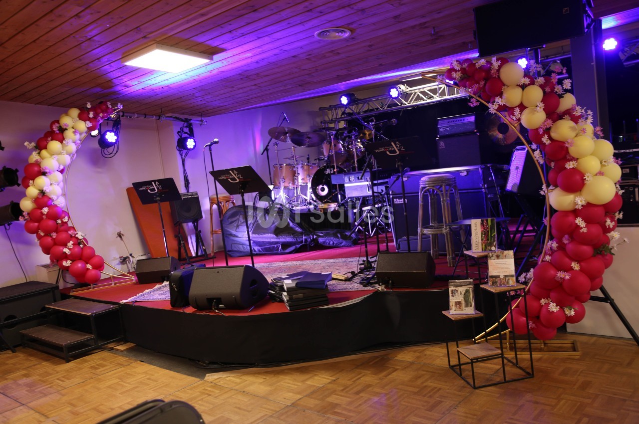 Scène décorée avec des ballons rouges et jaunes, équipée d'instruments de musique et de micros sous un éclairage violet.