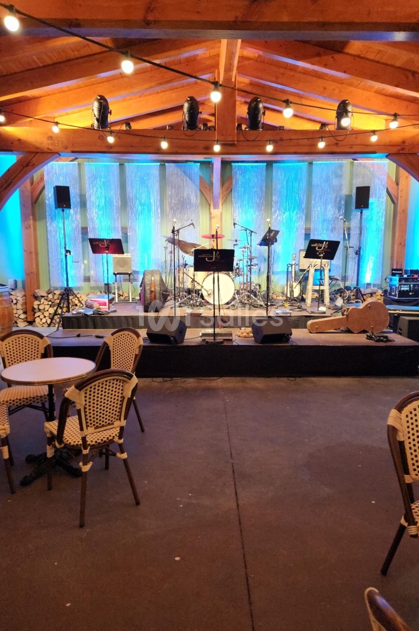 Scène de concert vide sous une structure en bois, éclairée par des lumières bleues, avec des tables et chaises au premier…