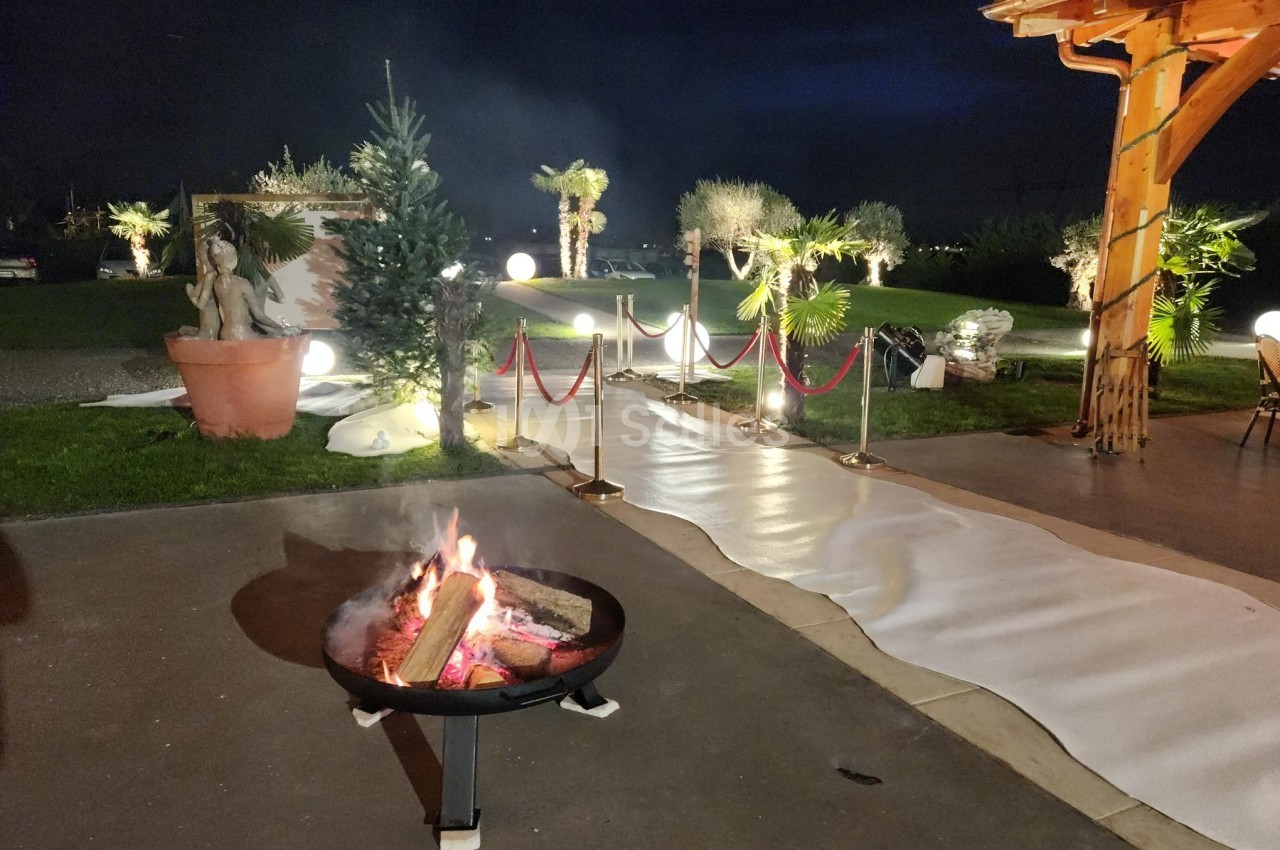 Brasero allumé sur une terrasse éclairée, avec un chemin blanc bordé de palmiers et de lampes dans un décor nocturne.
