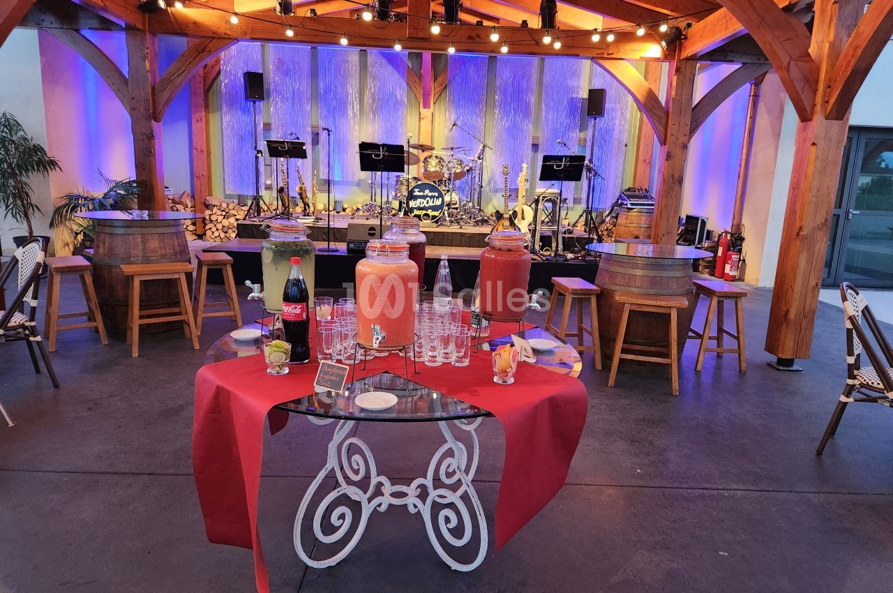 Buffet avec boissons et verres sur une table rouge, dans un espace couvert décoré avec éclairages et scène musicale en…