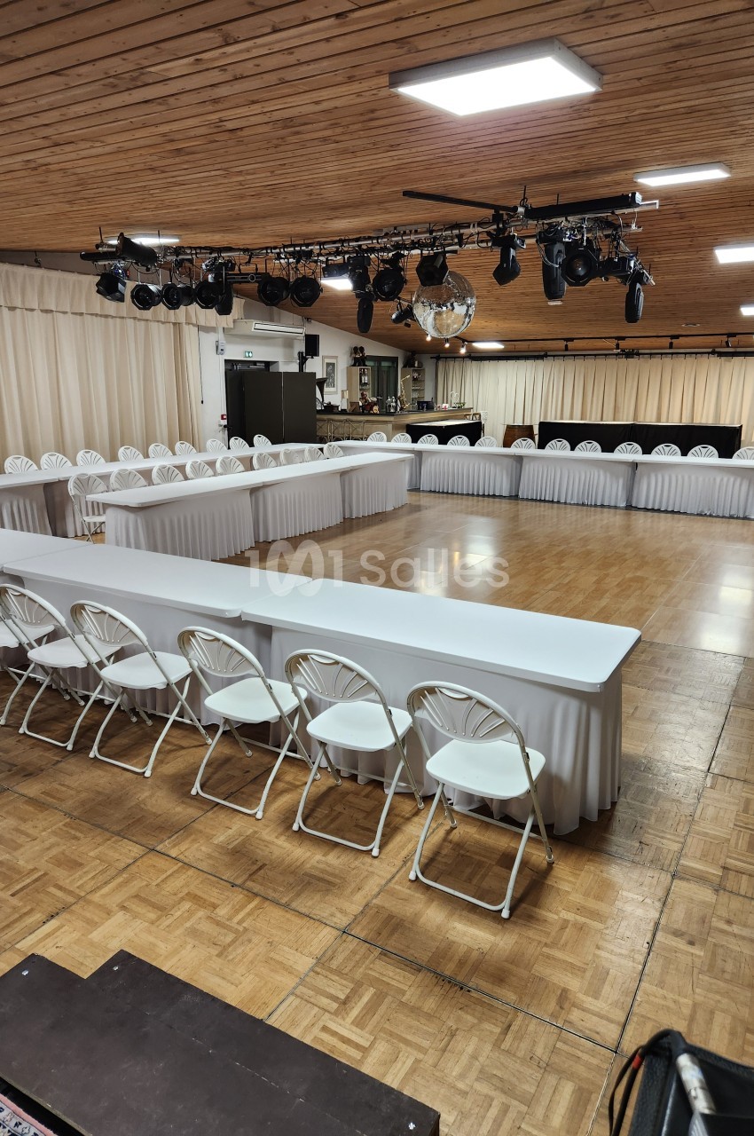 Salle de réception avec parquet, tables blanches disposées en U, chaises pliantes et éclairage suspendu.