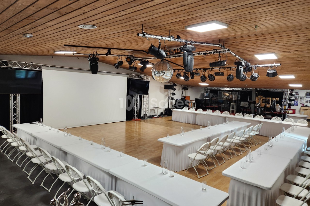 Salle de conférence avec tables en U, chaises blanches, écran de projection, éclairages suspendus et parquet au sol.