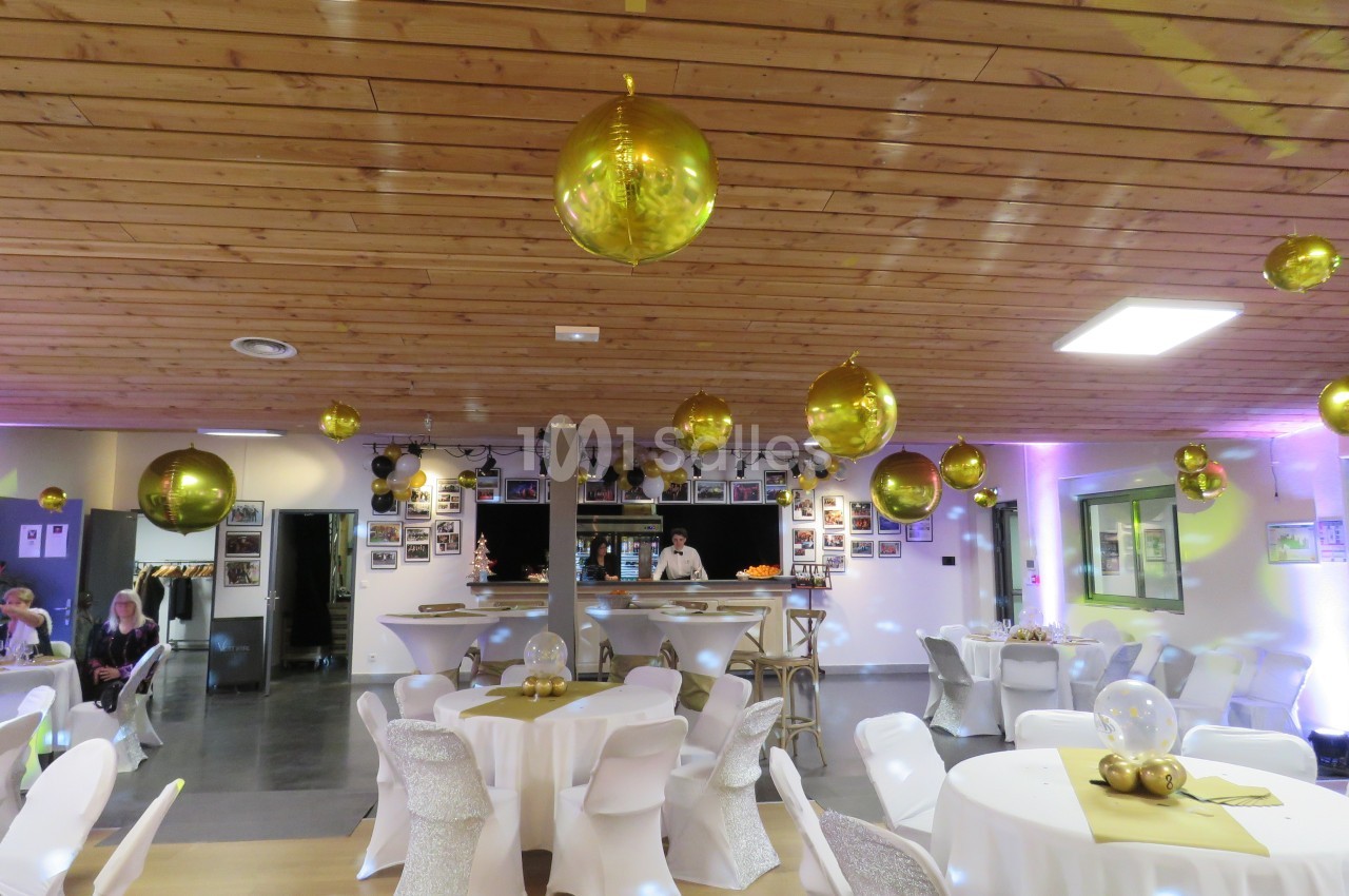 Salle décorée pour un événement avec des ballons dorés suspendus, des tables rondes dressées et un bar en arrière-plan.
