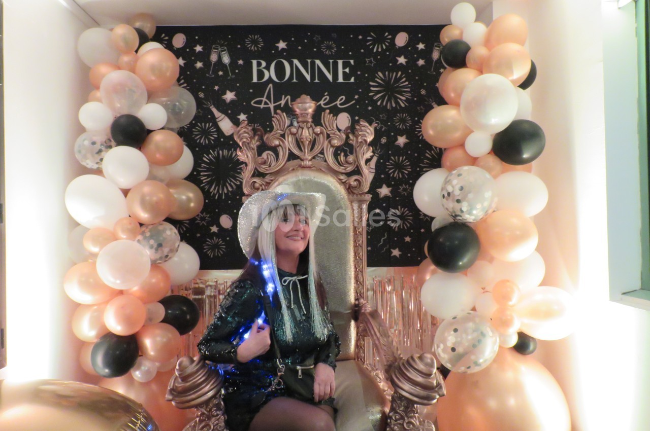 Femme souriante assise sur un trône décoré, entourée de ballons et d'une affiche ’Bonne Année’ festive en arrière-plan.