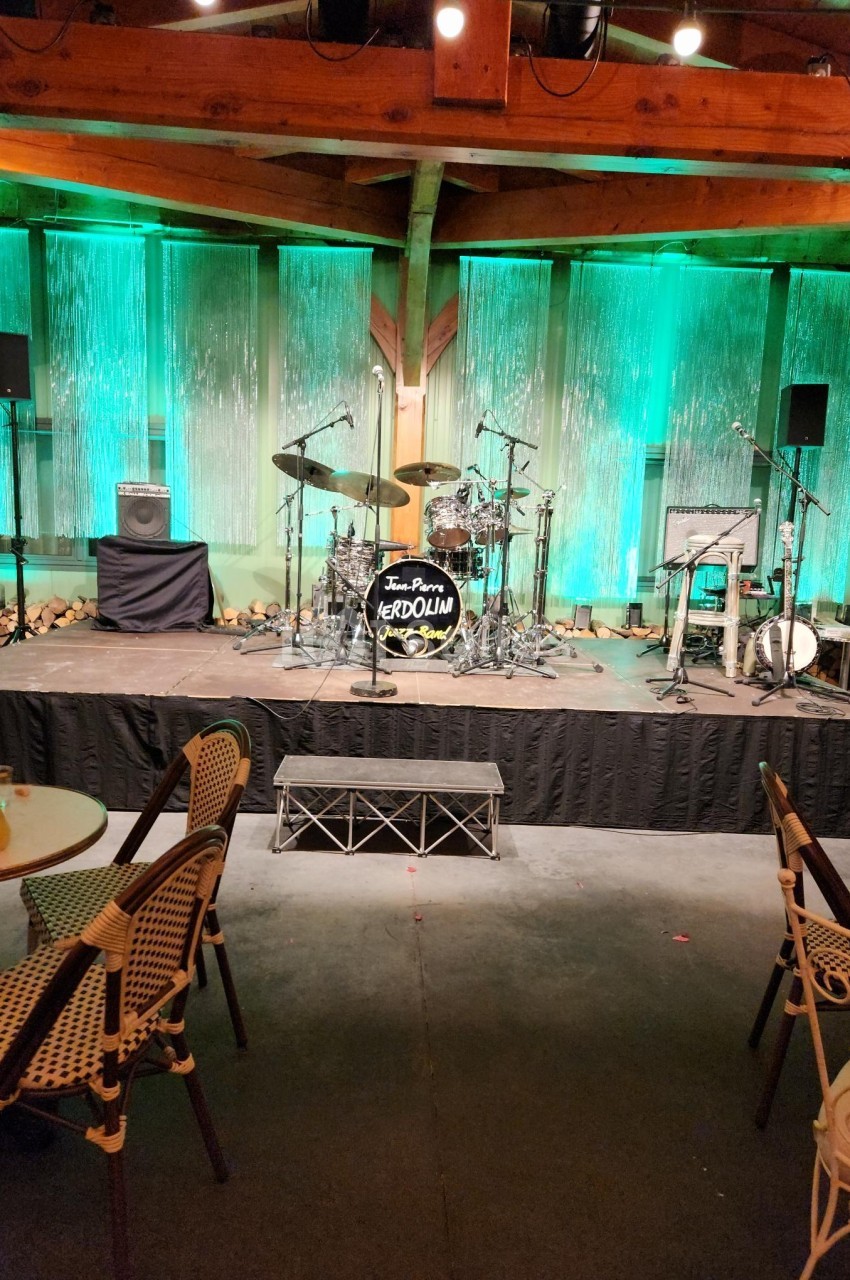 Scène vide avec une batterie, des amplificateurs et des éclairages verts, entourée de chaises et de tables en bois.
