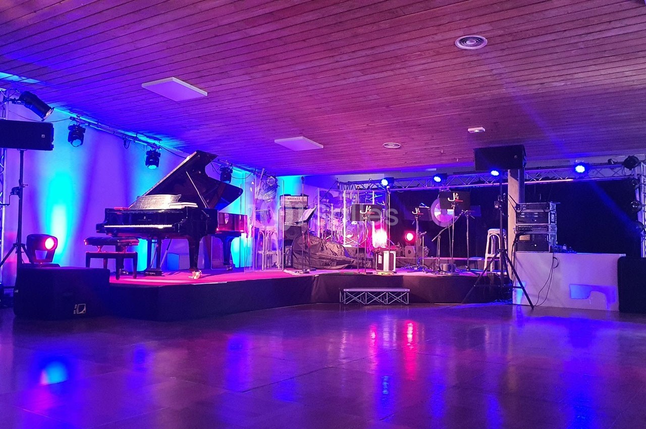 Salle de concert avec un piano à queue sur scène, éclairages bleus et rouges, et équipements audio en arrière-plan.