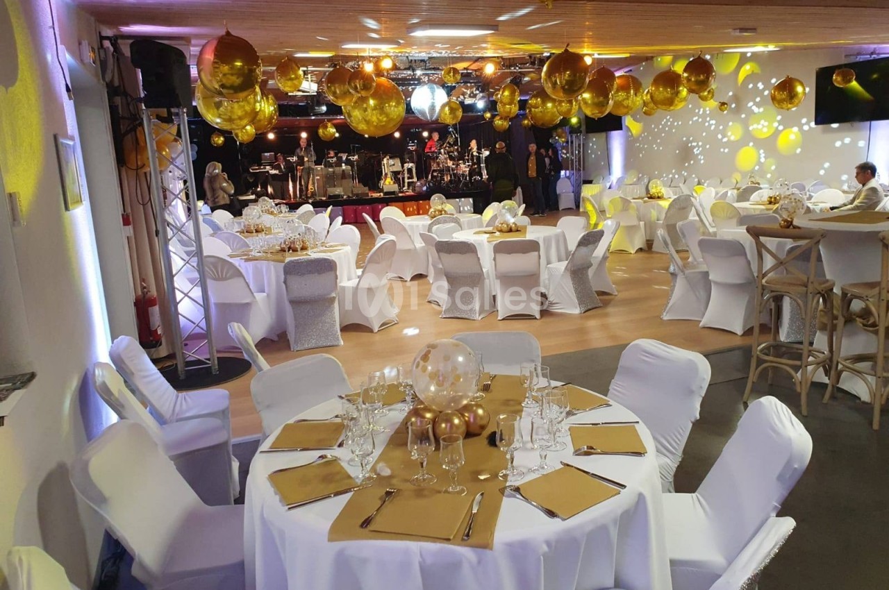 Salle décorée pour un événement avec tables rondes dressées, chaises blanches et ballons dorés suspendus.