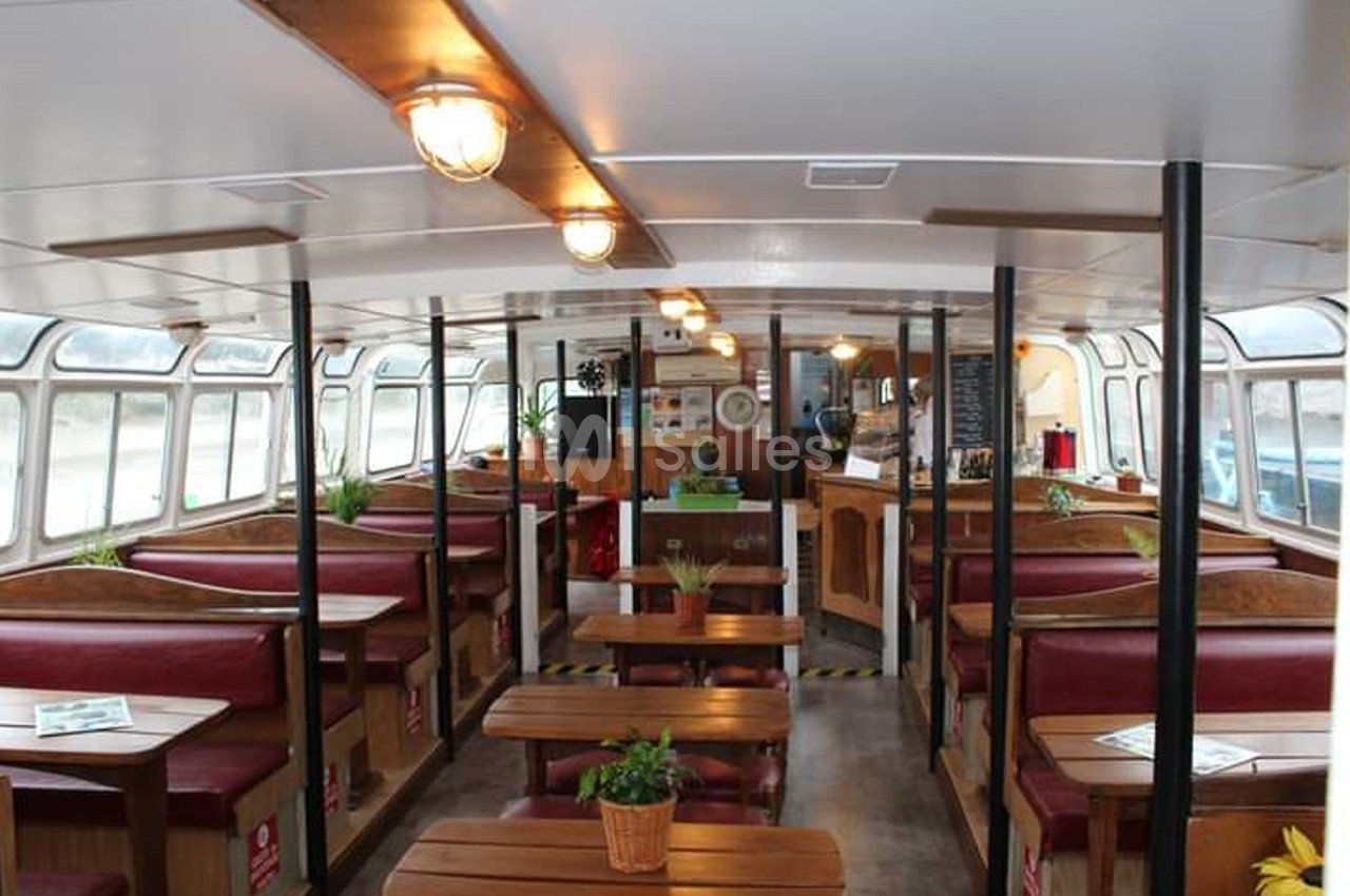 Intérieur d'un bateau aménagé en café avec banquettes rouges, tables en bois et comptoir au fond.