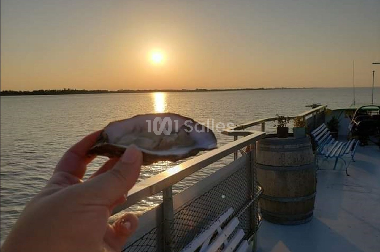 Une main tient une huître ouverte sur un bateau au coucher du soleil, avec vue sur l'eau calme et l'horizon.