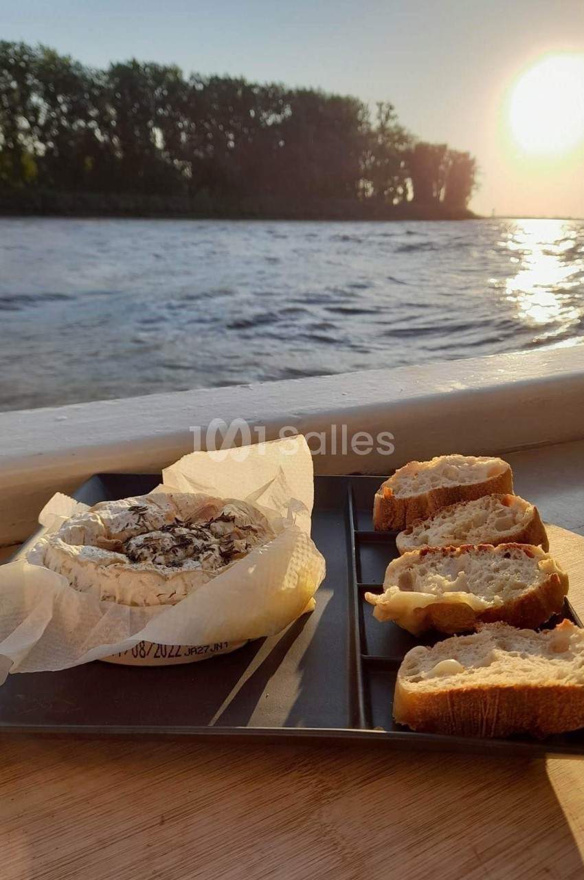 Fromage coulant et tranches de pain sur un plateau noir, posés près d'une fenêtre avec vue sur un lac au coucher du soleil.