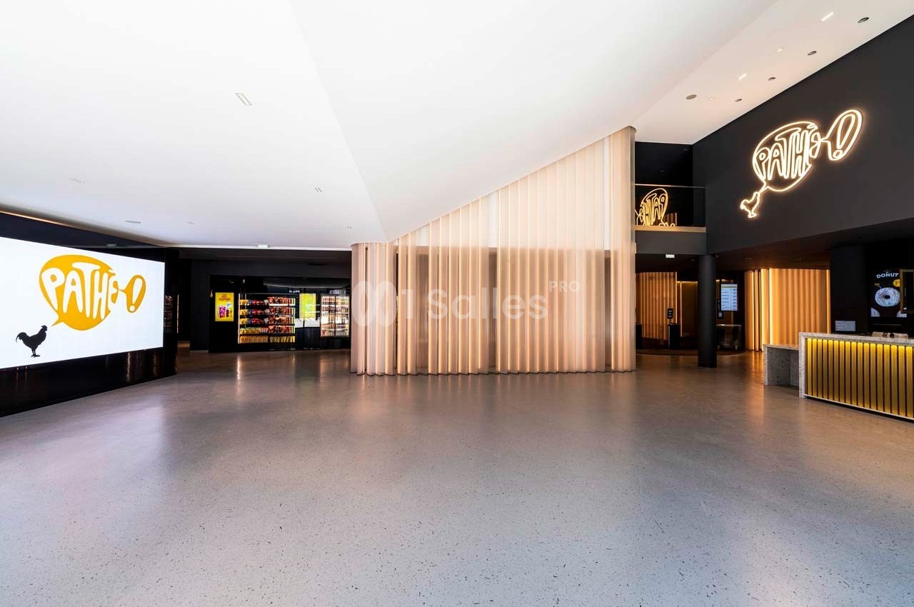 Hall d'entrée d'un cinéma avec un sol en béton, des panneaux lumineux et un comptoir d'accueil éclairé.