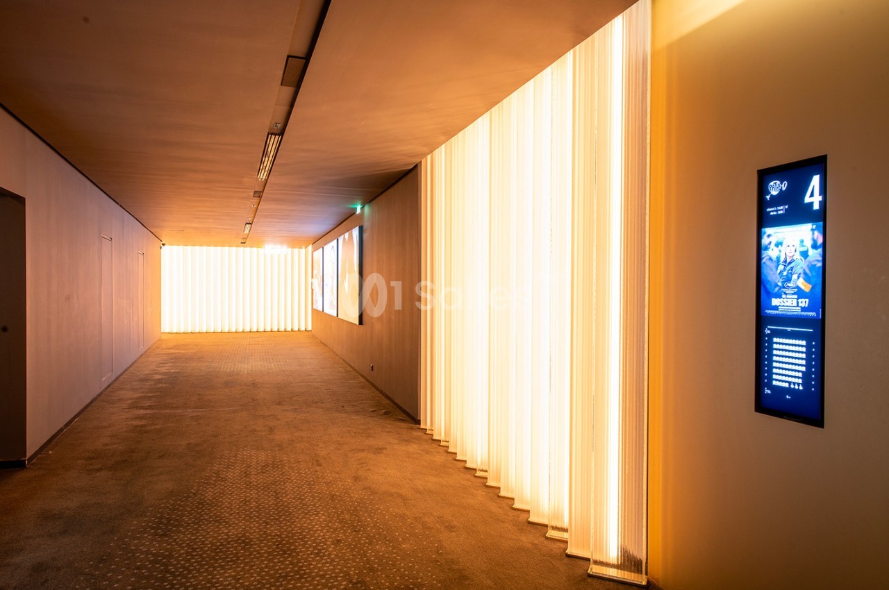Couloir moderne avec éclairage mural lumineux et écran numérique affichant des informations sur la droite.