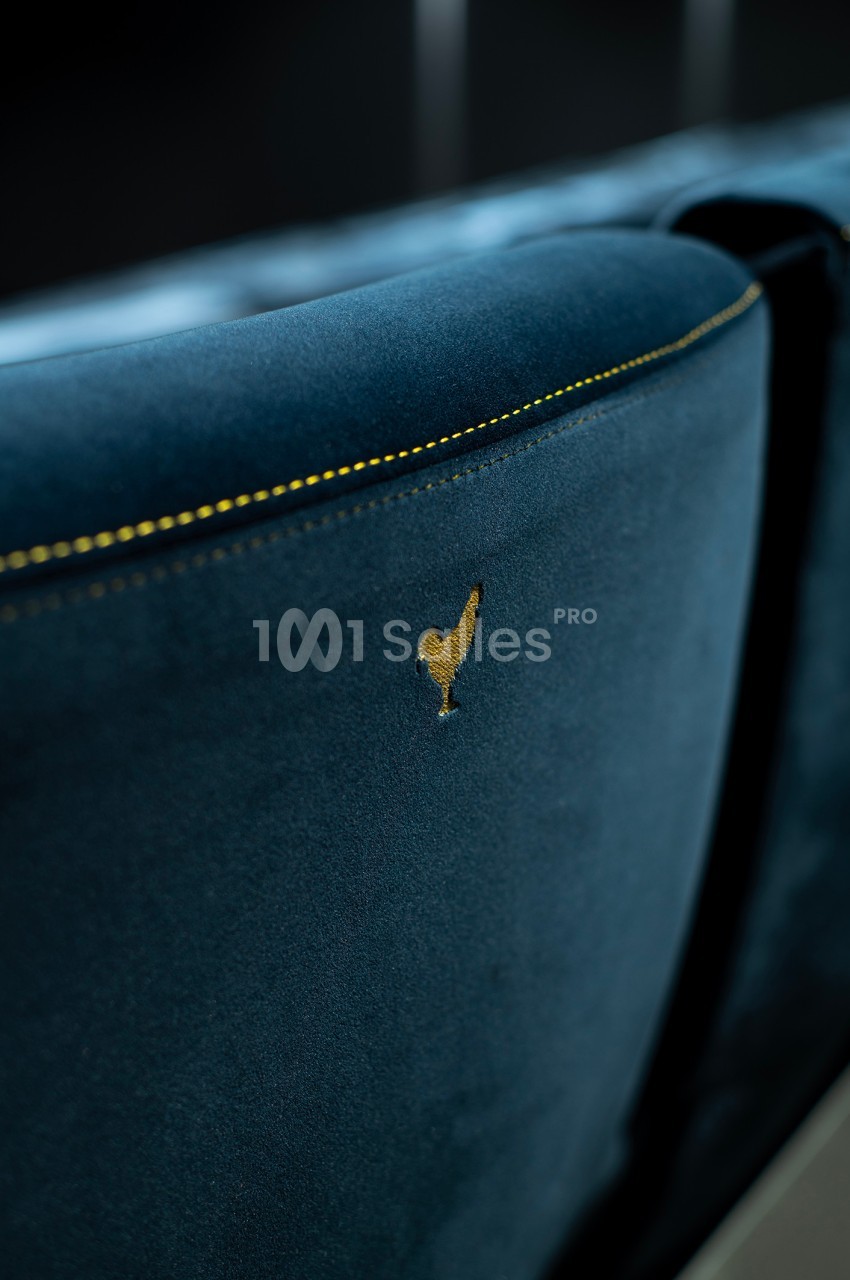 Dossier de fauteuil en velours bleu avec une broderie dorée en forme de coq, éclairé par une lumière douce.