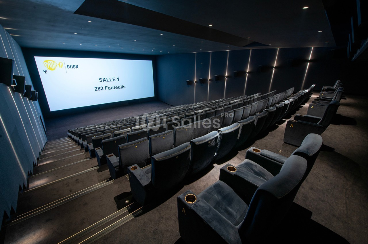 Salle de cinéma moderne avec rangées de fauteuils en velours gris et grand écran affichant ’Salle 1, 282 fauteuils’.