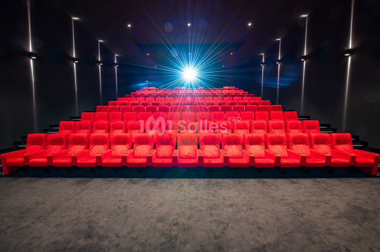 Salle de cinéma vide avec des rangées de fauteuils rouges et un projecteur allumé au fond.