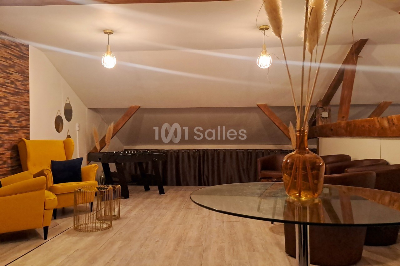 Espace salon mansardé avec fauteuils, table en verre, baby-foot et décoration moderne sous poutres apparentes.