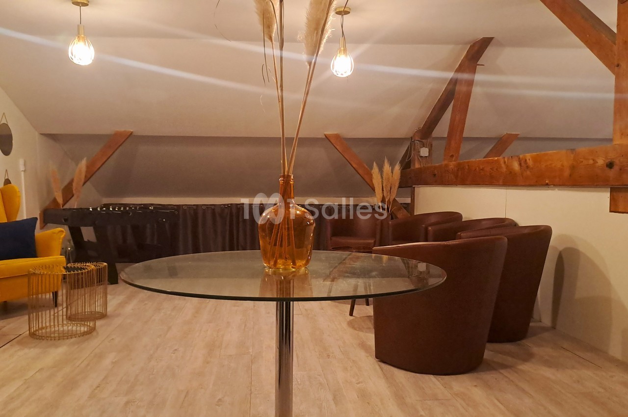 Salle mansardée avec table en verre, fauteuils en cuir marron, fauteuil jaune et décoration minimaliste.