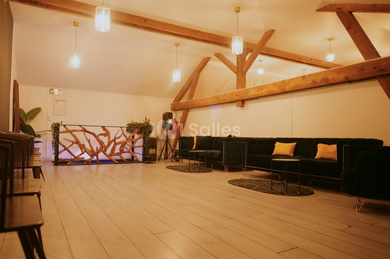 Salle lumineuse avec poutres apparentes, canapés bleus, coussins jaunes, plantes et sol en bois clair.
