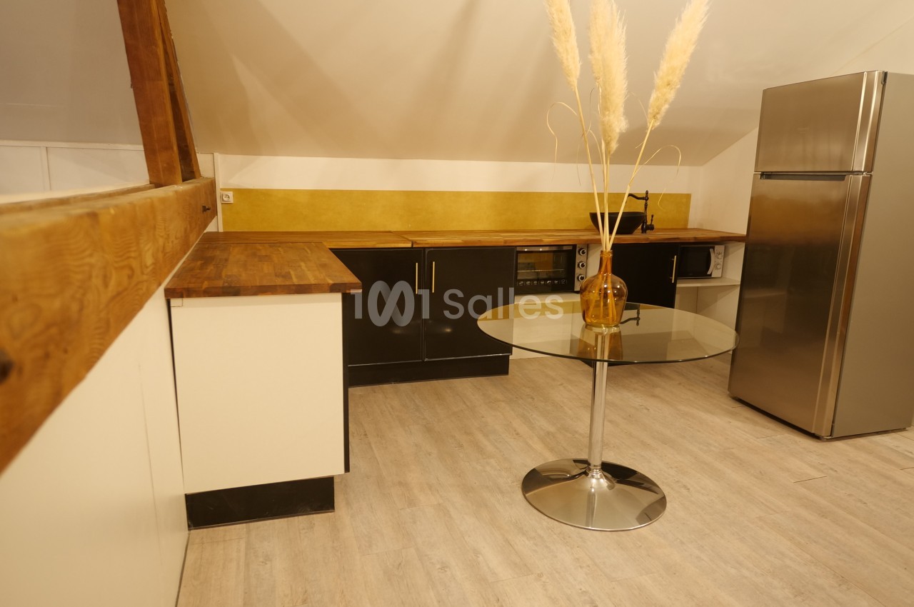 Cuisine sous combles avec meubles noirs, plan de travail en bois, table ronde en verre et réfrigérateur en inox.