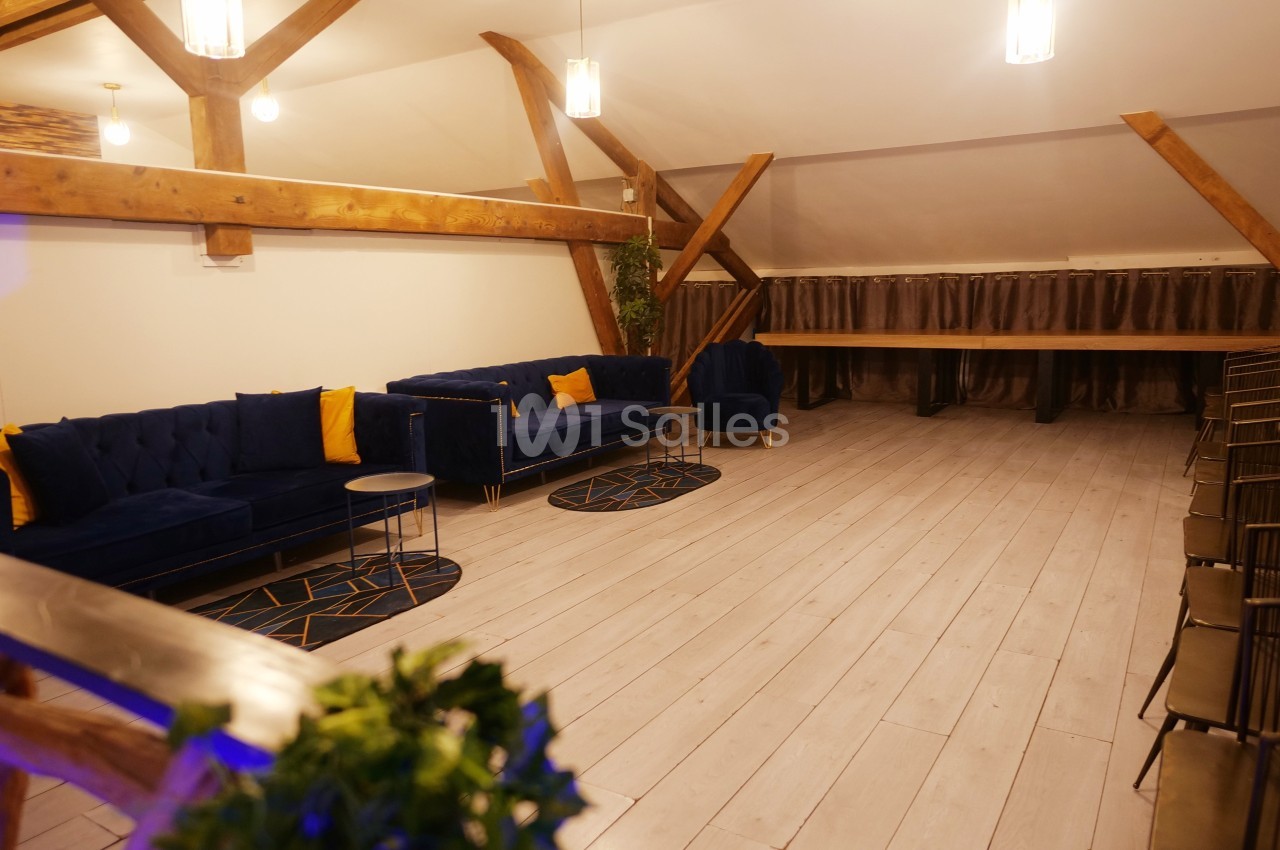 Salle lumineuse avec parquet clair, canapés bleus, coussins jaunes, tables basses et poutres apparentes en bois.