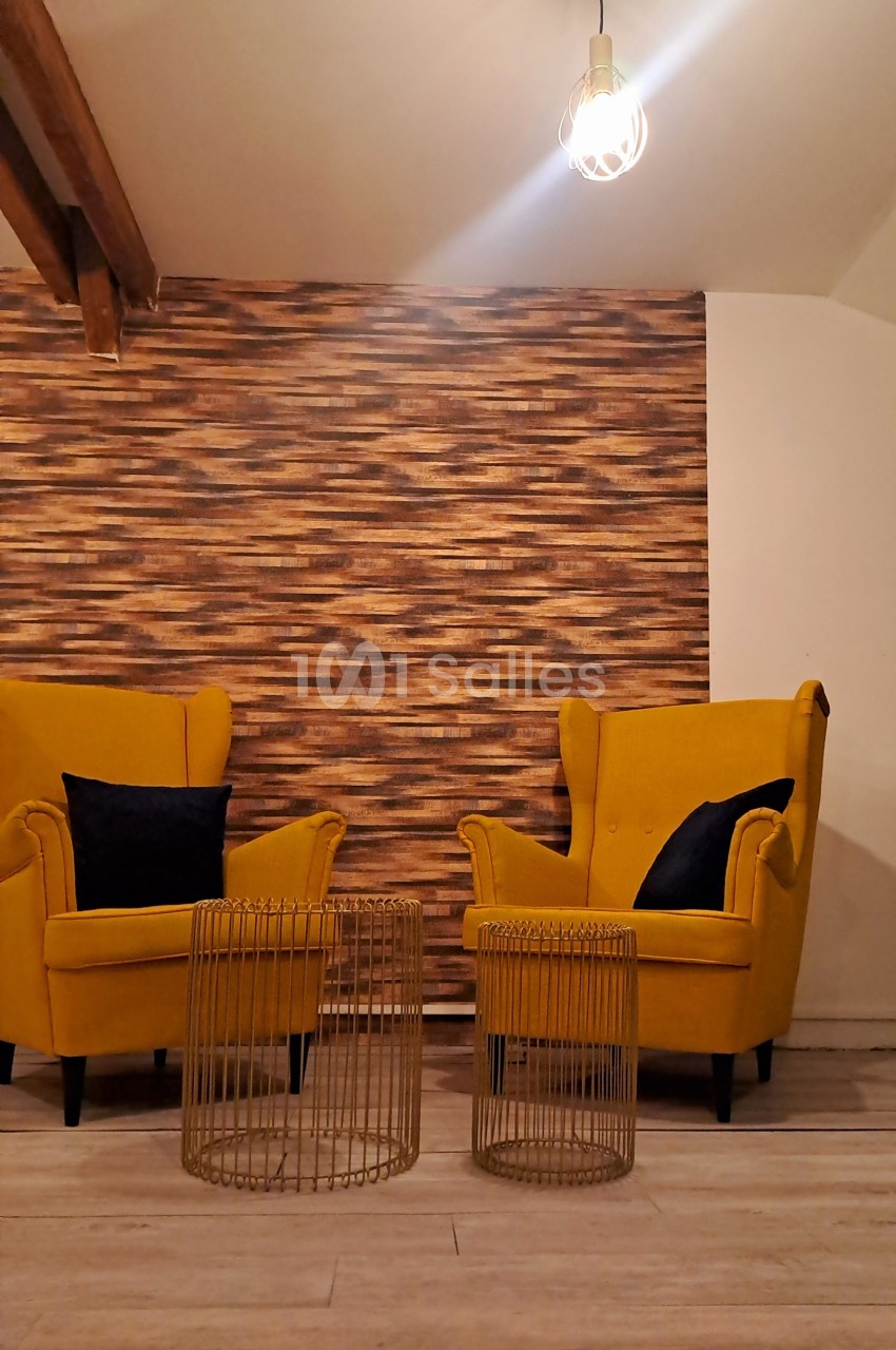 Deux fauteuils jaunes avec coussins noirs entourent deux tables basses dorées, devant un mur texturé en bois.