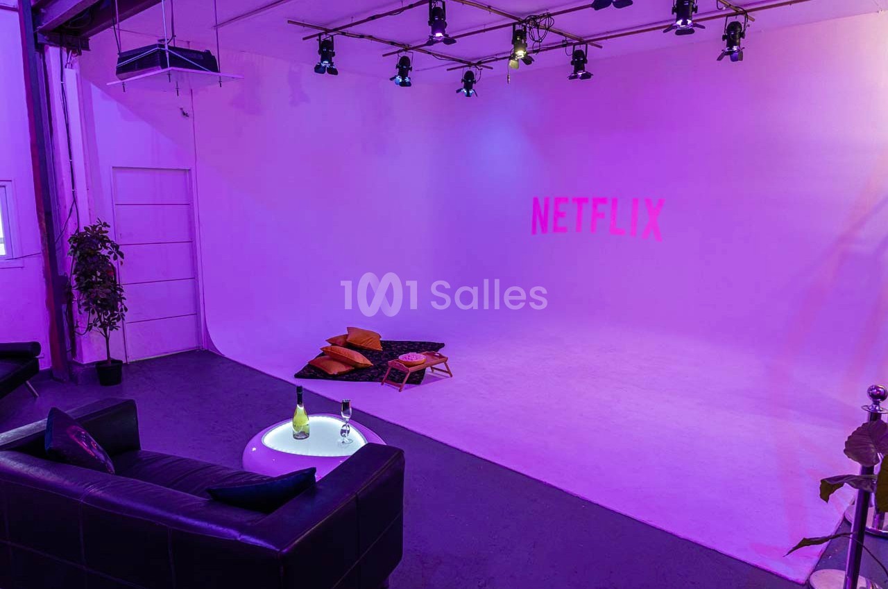 Studio éclairé en violet avec un canapé, une table basse et des coussins au sol, logo Netflix projeté sur le mur.