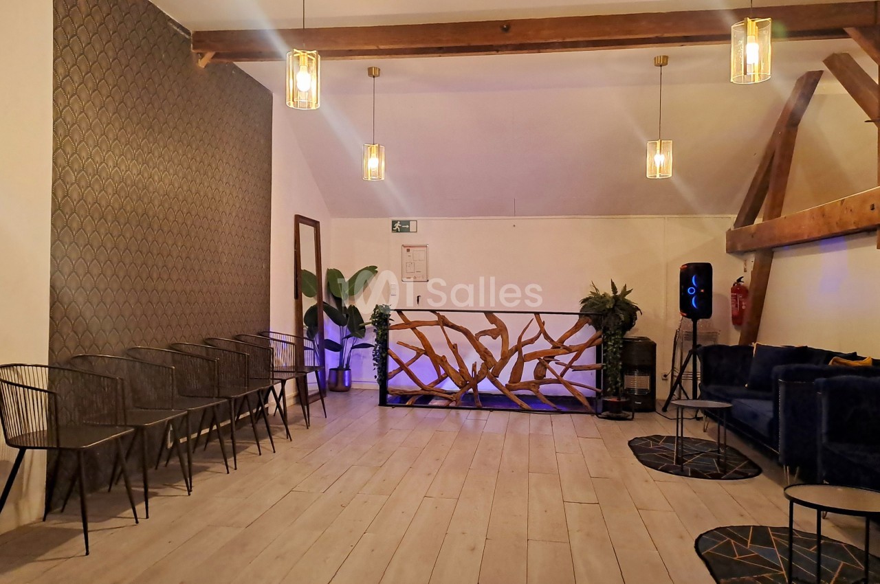 Salle lumineuse avec parquet clair, chaises alignées, canapé bleu, plantes et balustrade décorative en bois sculpté.
