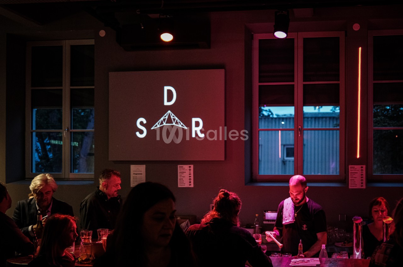 Un barman sert des boissons dans un bar faiblement éclairé avec un logo projeté sur un mur en arrière-plan.