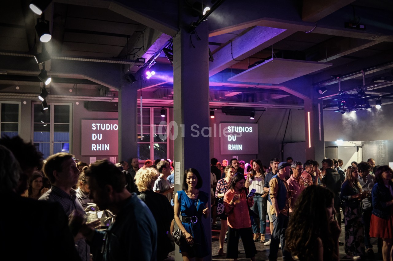 Salle animée avec des personnes debout discutant lors d'un événement, éclairée par des lumières colorées.
