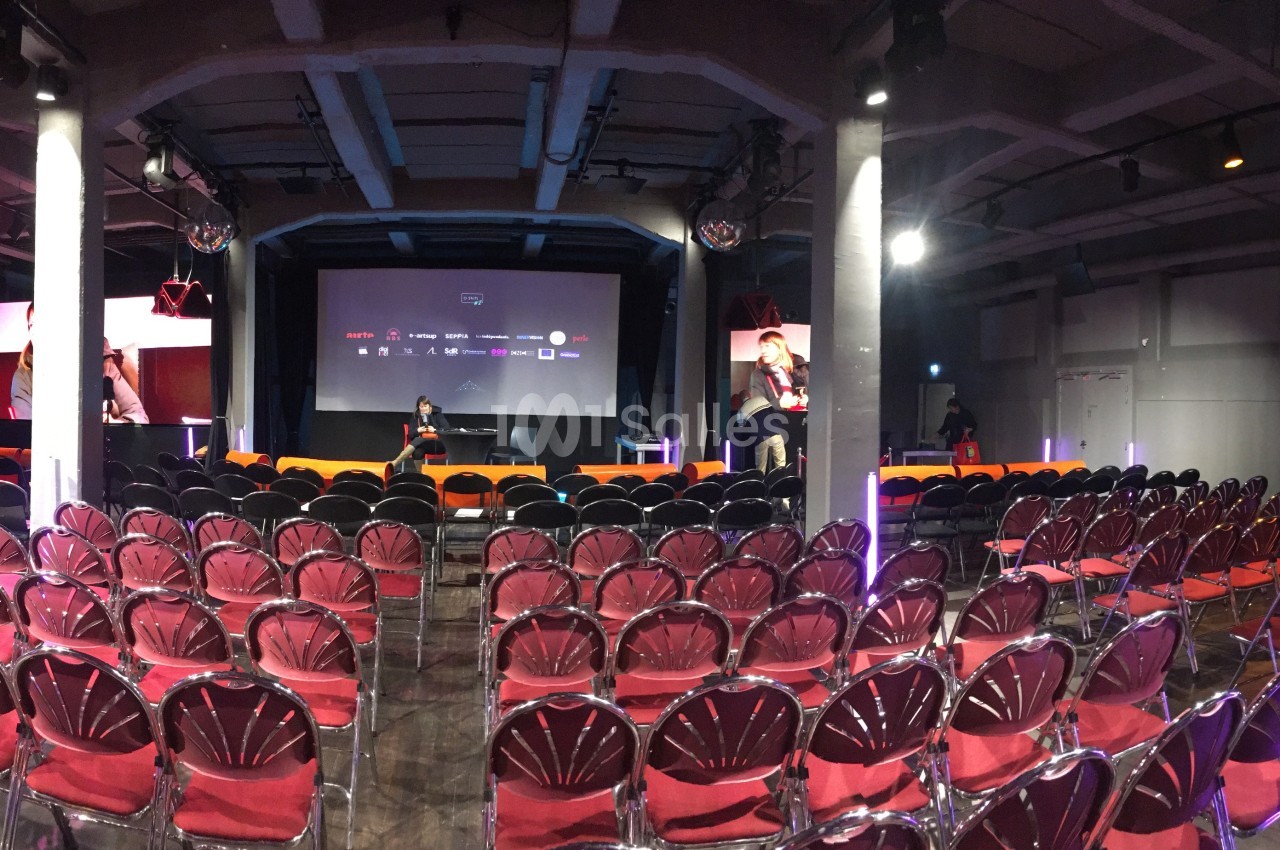 Salle de conférence vide avec rangées de chaises rouges, éclairage de scène et écran de projection en arrière-plan.