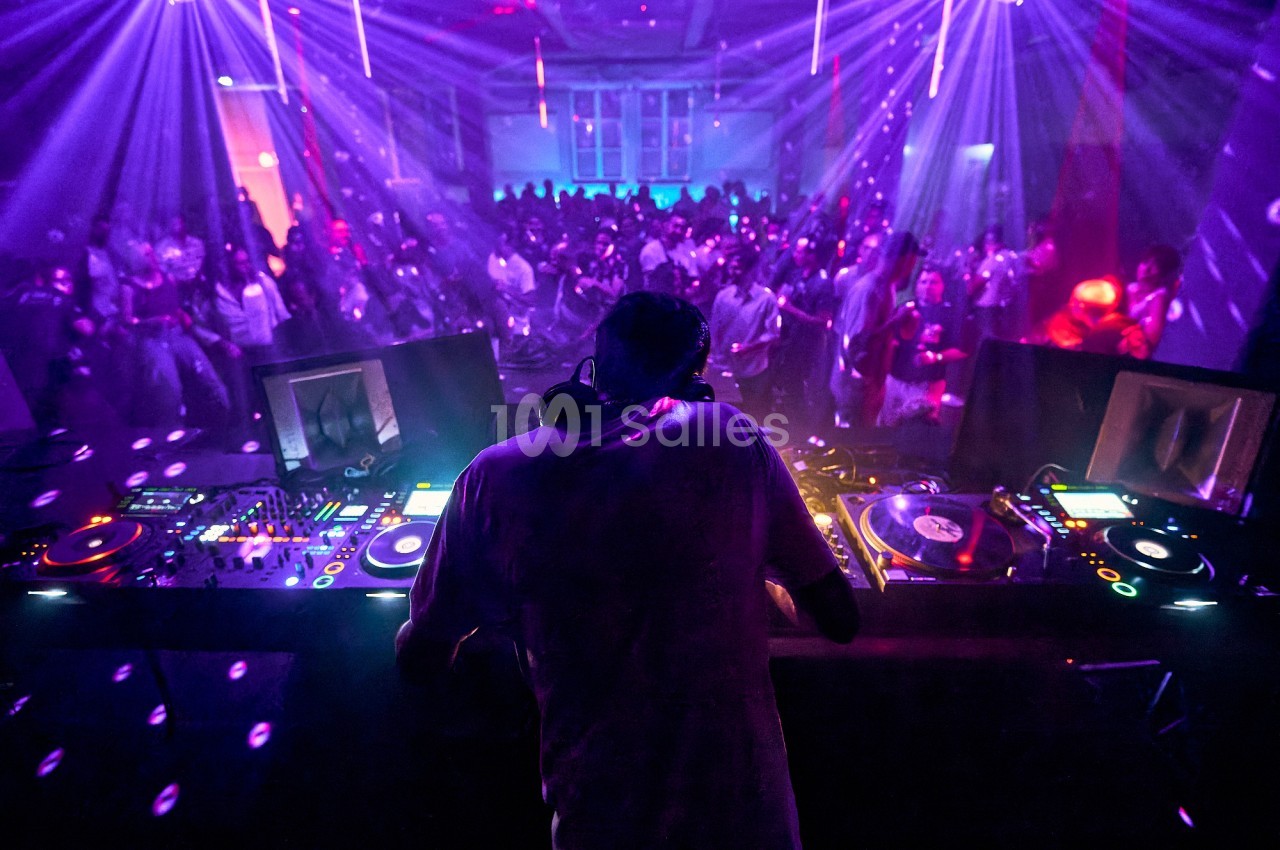 Un DJ mixe sur une scène éclairée par des lumières colorées, face à une foule dansante dans une salle sombre.