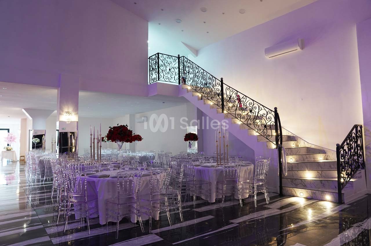 Salle de réception élégante avec tables dressées, chaises transparentes et escalier décoratif éclairé.