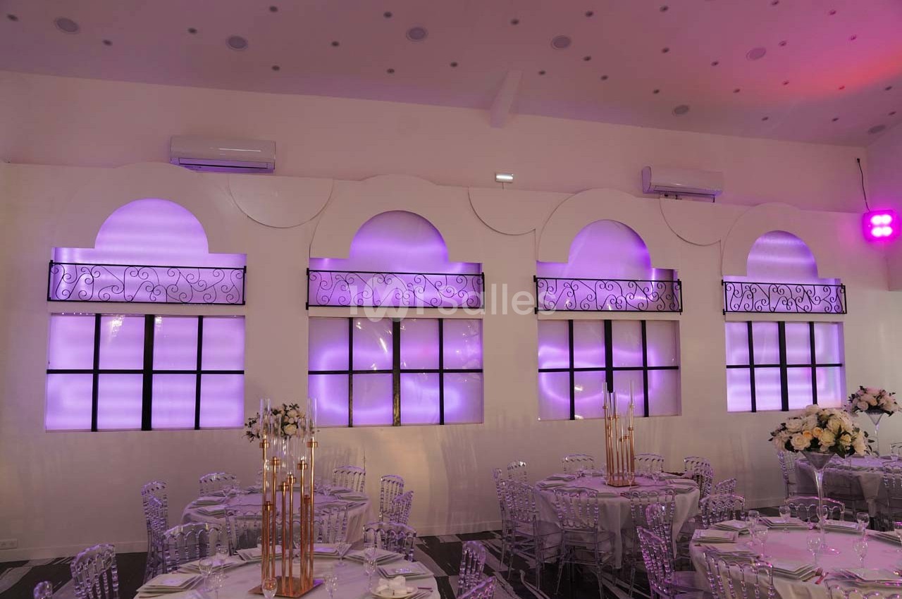 Salle de réception avec tables dressées, chaises transparentes et éclairage violet diffus sur des fenêtres décoratives.