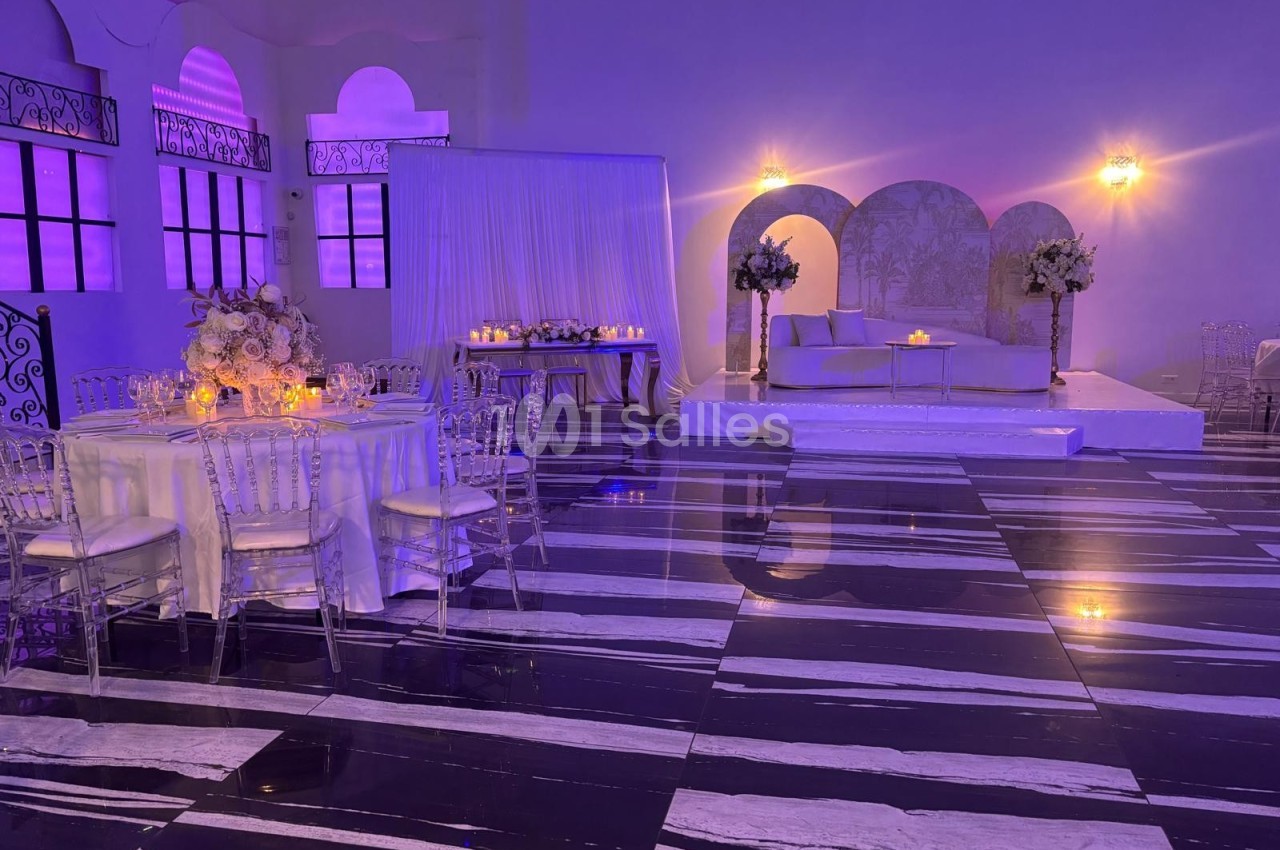 Salle de réception élégante avec tables décorées, éclairage violet et espace lounge sur une estrade.