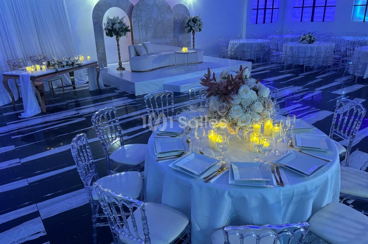 Salle de réception élégante avec tables décorées de fleurs et bougies, éclairage bleu tamisé et estrade en arrière-plan.