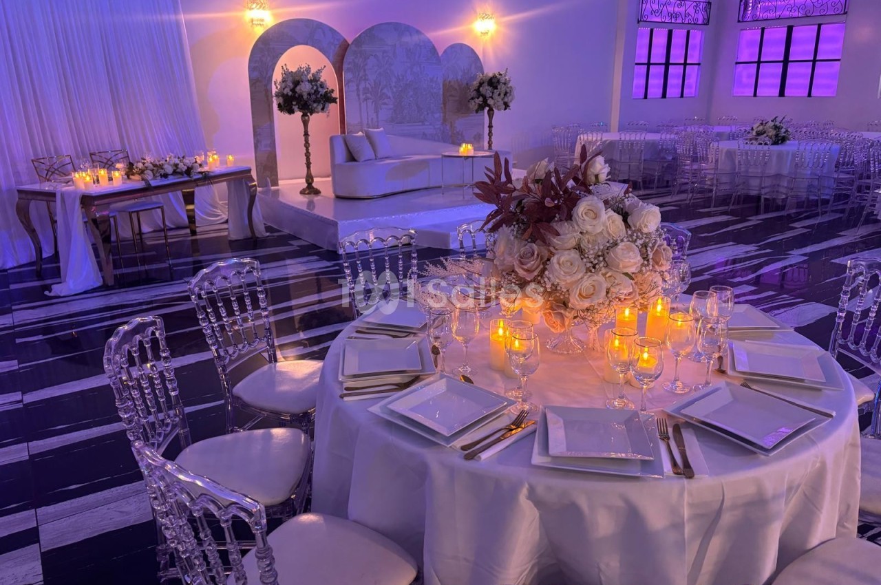 Salle de réception élégante avec tables décorées de bougies et fleurs, chaises transparentes et éclairage tamisé.