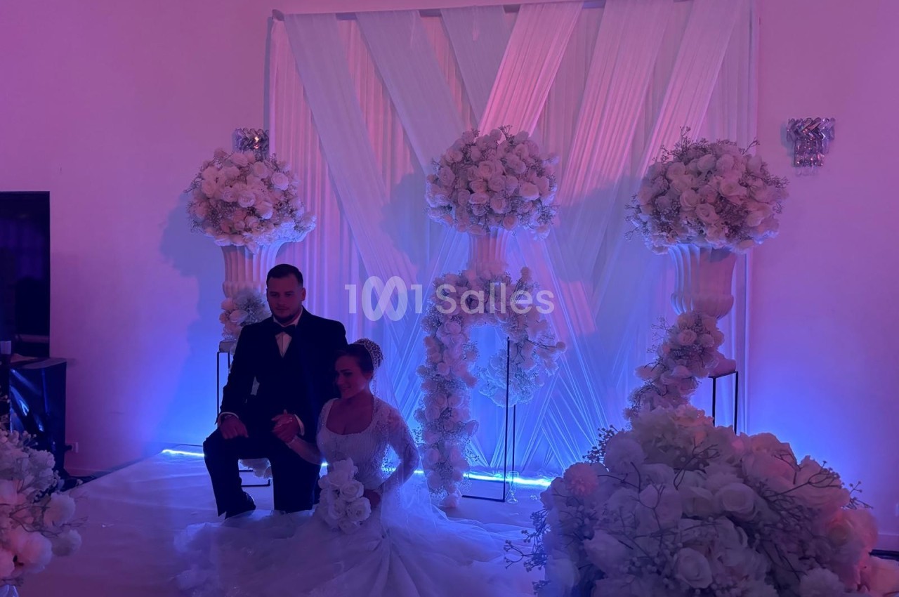 Un couple en tenue de mariage pose devant une décoration florale blanche éclairée par une lumière violette.