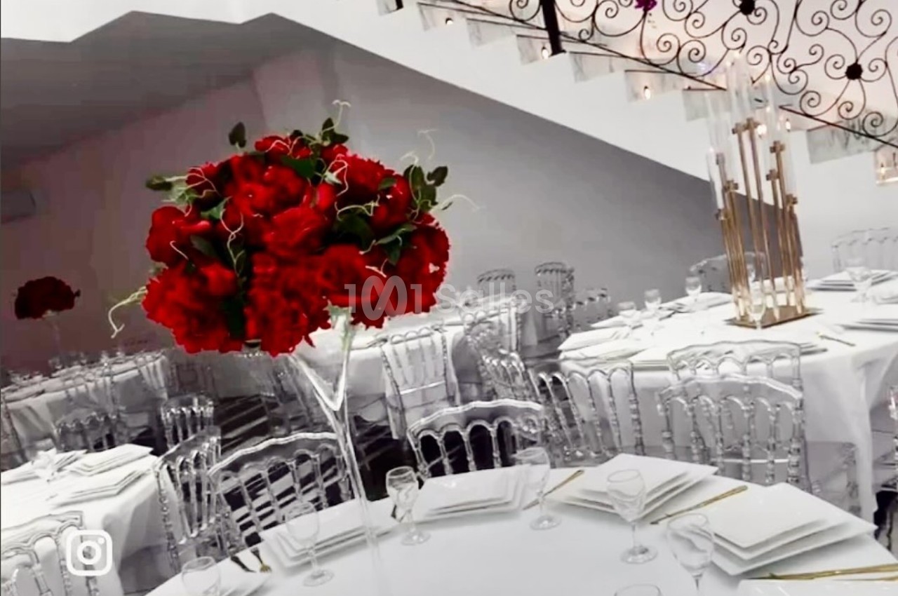 Salle de réception élégante avec tables dressées, centre de table floral rouge et escalier à rambarde en fer forgé.