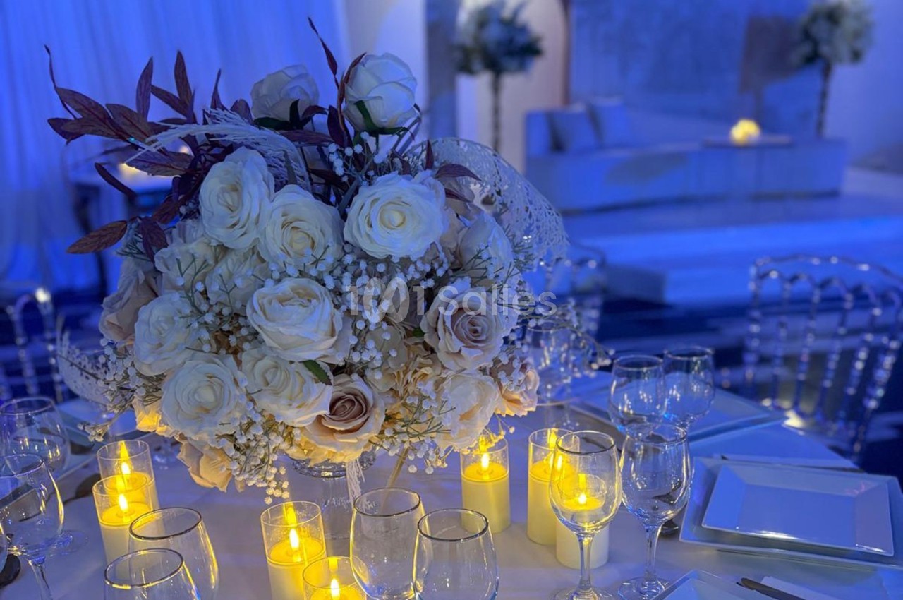 Centre de table avec roses blanches et bougies allumées, entouré de verres et d'assiettes dans une ambiance feutrée.