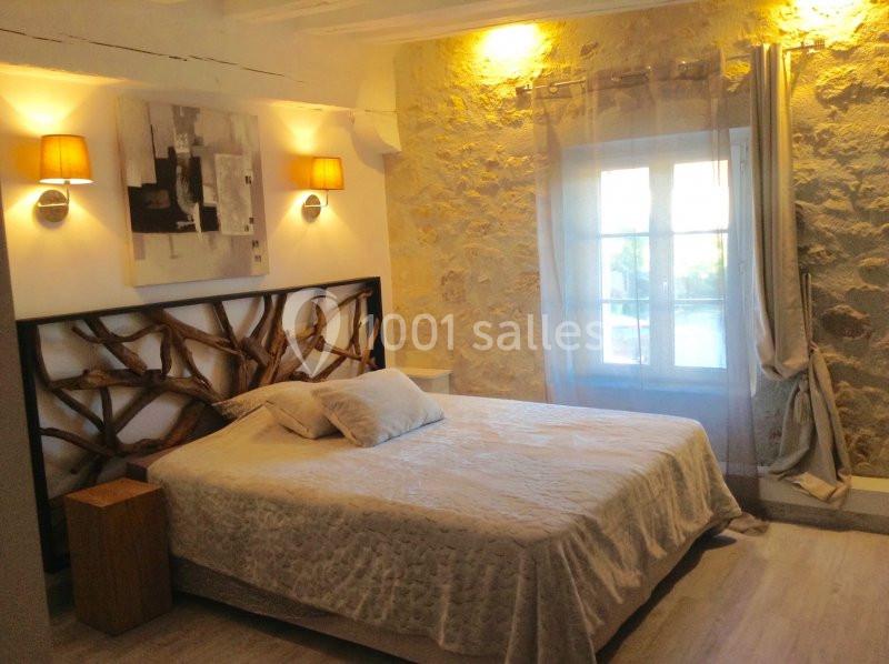 Chambre avec lit double, mur en pierre apparente, éclairage doux, tableau moderne et fenêtre avec rideaux translucides.